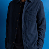 A man wearing a dark navy blue textured button-up shirt over a black t-shirt, set against a solid blue studio background.