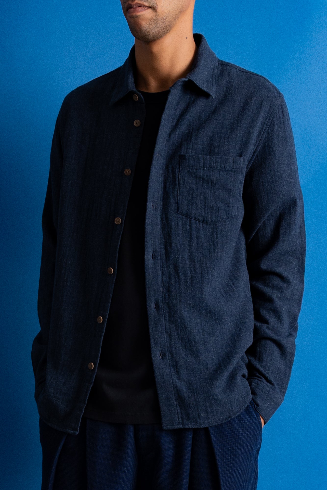 A man wearing a dark navy blue textured button-up shirt over a black t-shirt, set against a solid blue studio background.