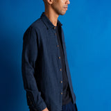 A man wearing a dark navy blue textured button-up shirt over a black t-shirt and navy pants, standing against a solid blue studio background.