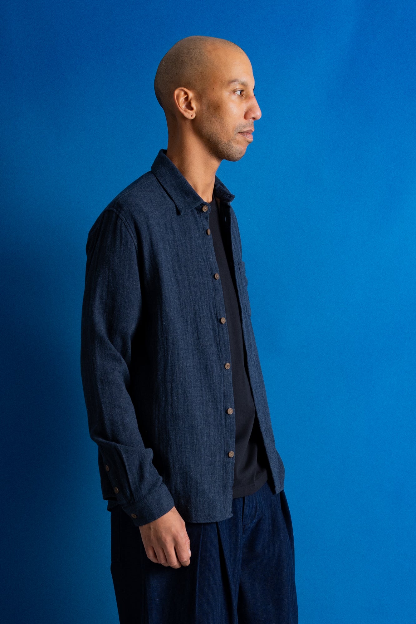 A man wearing a dark navy blue textured button-up shirt over a black t-shirt and navy pants, standing against a solid blue studio background.