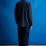 A man wearing a dark navy blue textured button-up shirt over a black t-shirt and navy pants, standing against a solid blue studio background.