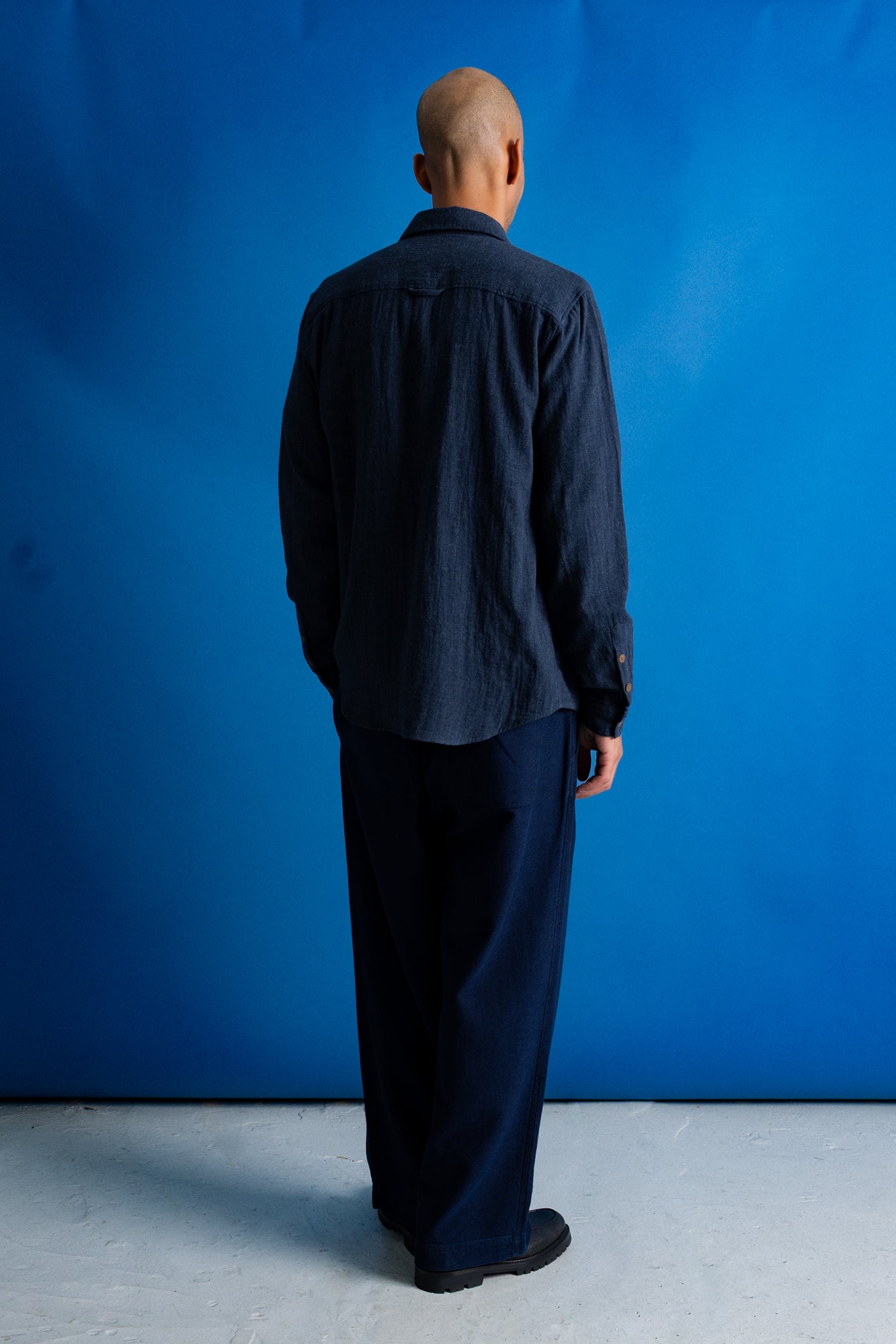 A man wearing a dark navy blue textured button-up shirt over a black t-shirt and navy pants, standing against a solid blue studio background.