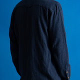 A man viewed from the back wearing a dark navy blue linen button-up shirt and matching navy trousers, set against a solid blue studio background.