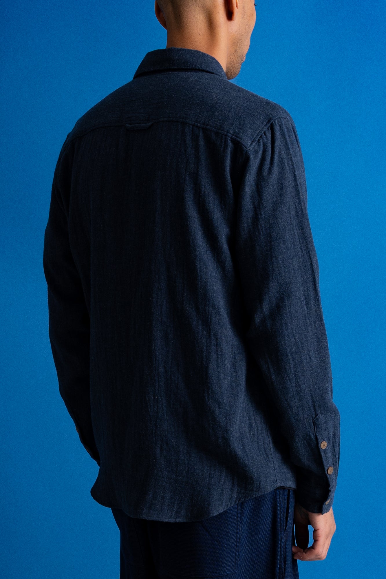 A man viewed from the back wearing a dark navy blue linen button-up shirt and matching navy trousers, set against a solid blue studio background.