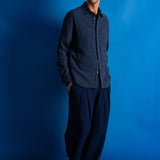 A man wearing a dark navy blue textured button-up shirt and matching dark navy trousers, standing against a vibrant blue studio backdrop.