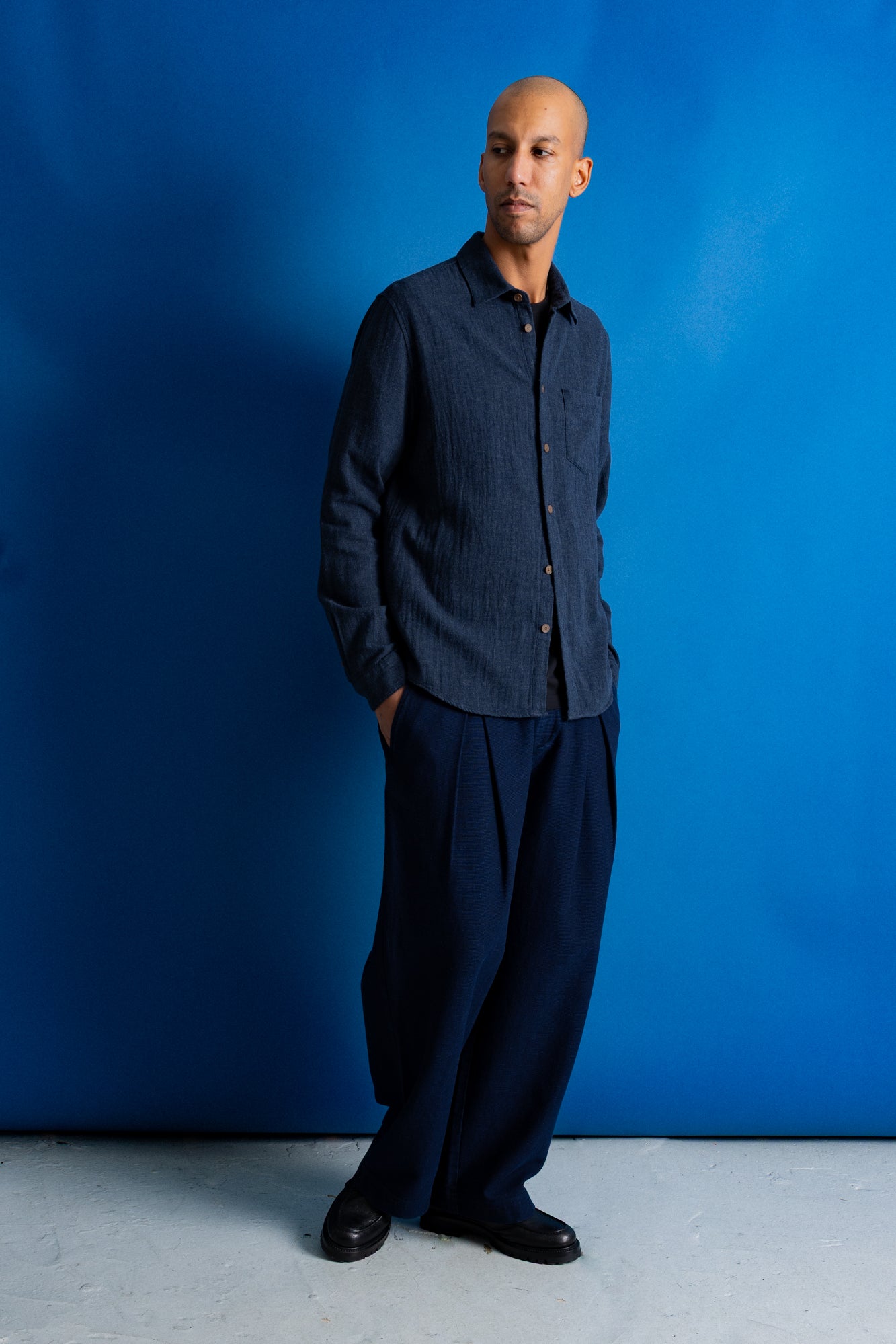 A man wearing a dark navy blue textured button-up shirt and matching dark navy trousers, standing against a vibrant blue studio backdrop.