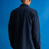 A man wearing a dark navy blue textured button-up shirt over a black t-shirt and navy pants, standing against a solid blue studio background.