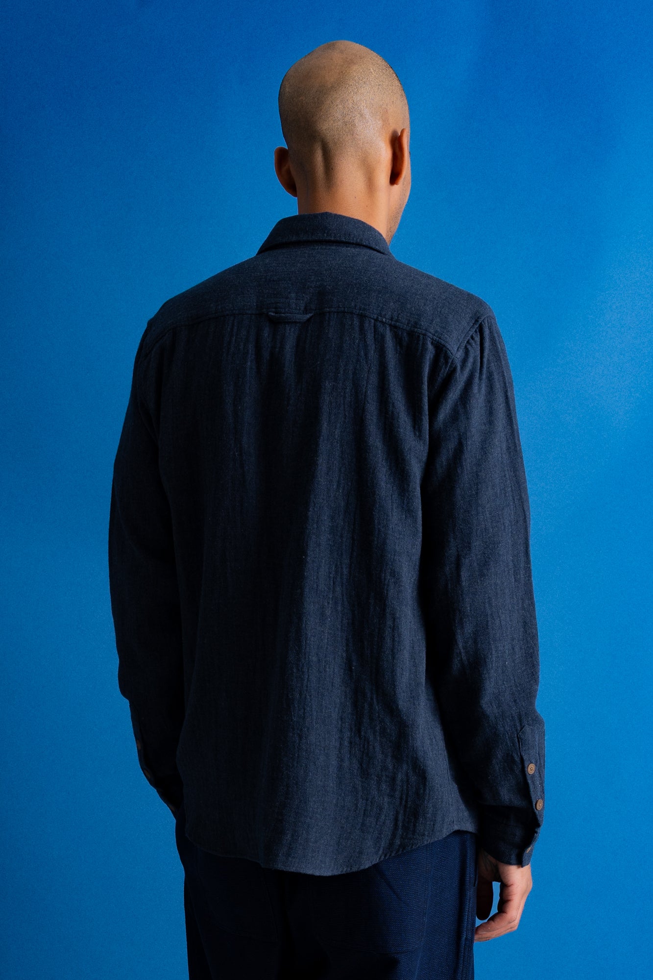 A man wearing a dark navy blue textured button-up shirt over a black t-shirt and navy pants, standing against a solid blue studio background.