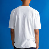 A man viewed from behind wearing a plain white short-sleeve t-shirt and dark pants, standing against a solid blue studio background.
