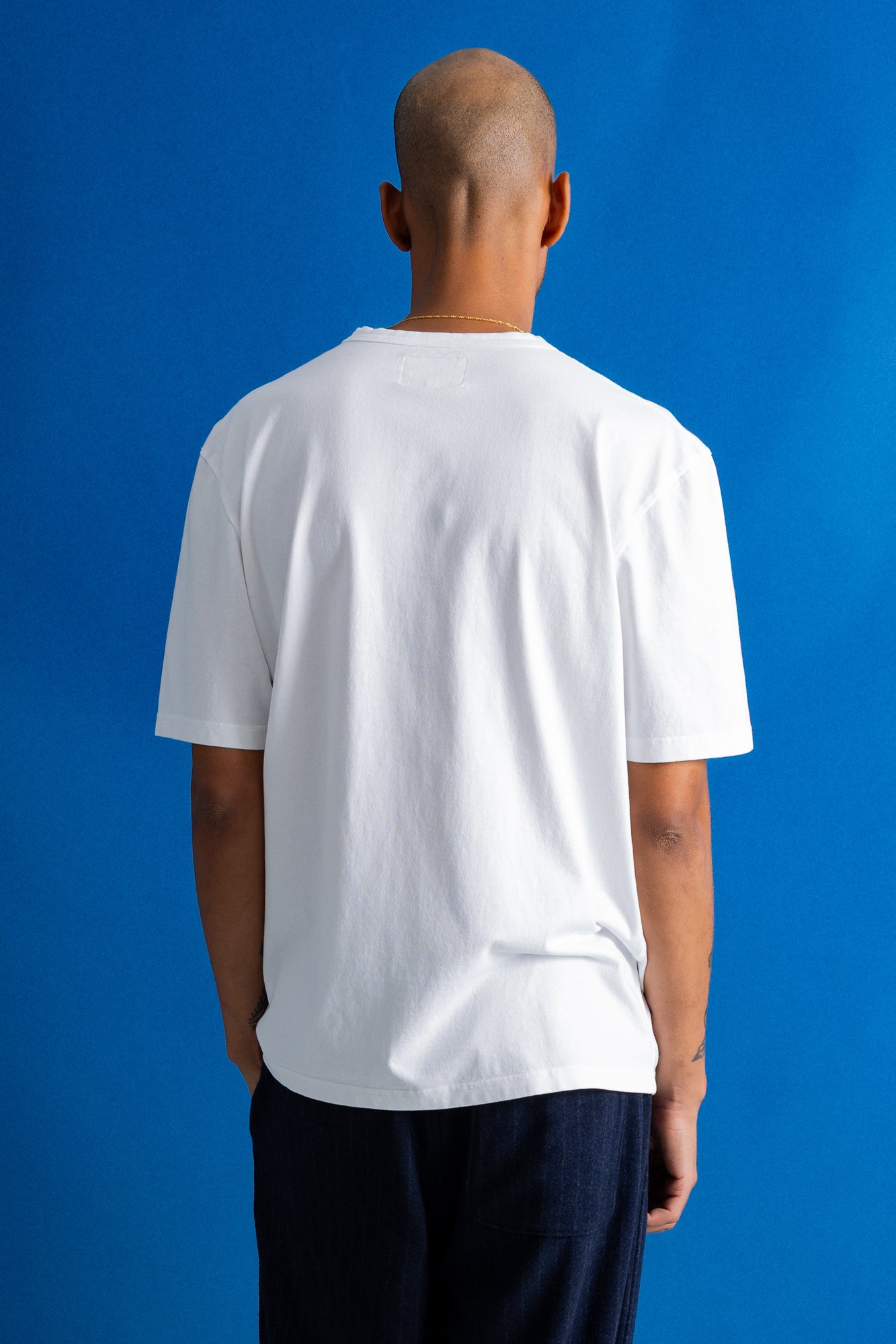 A man viewed from behind wearing a plain white short-sleeve t-shirt and dark pants, standing against a solid blue studio background.