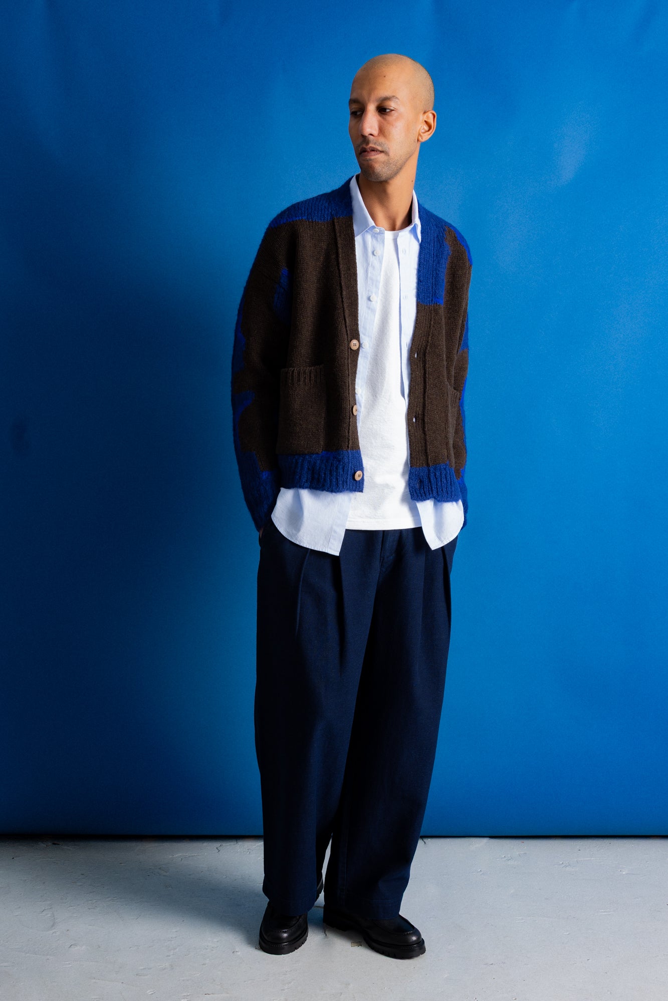 A man wearing a brown and blue knit cardigan over a white button-up shirt, paired with dark navy trousers and black shoes, standing against a solid blue studio backdrop.