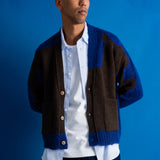 A man wearing a brown and blue color-block knit cardigan over a white button-down shirt, posed against a solid blue studio background.