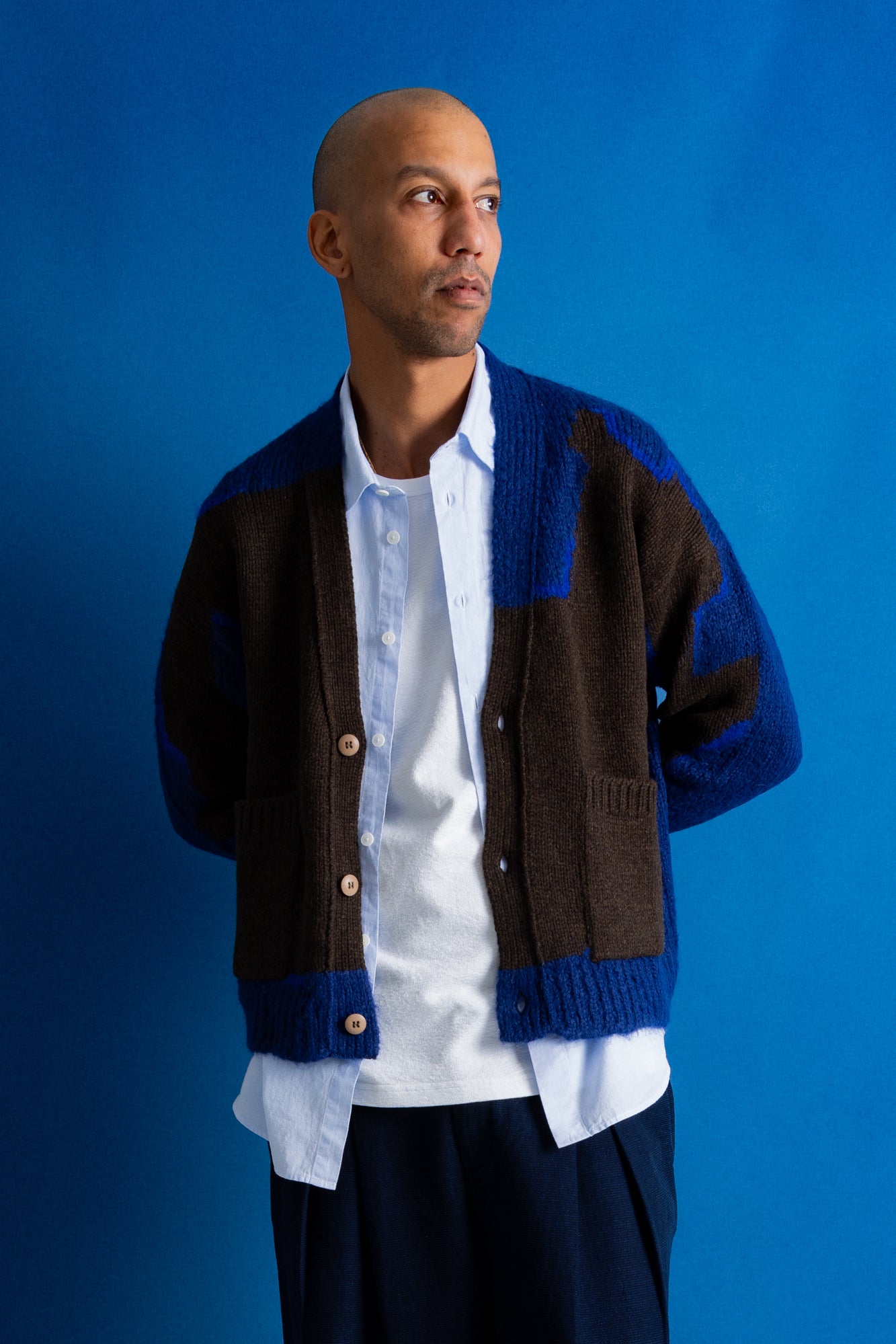 A man wearing a brown and blue color-block knit cardigan over a white button-down shirt, posed against a solid blue studio background.