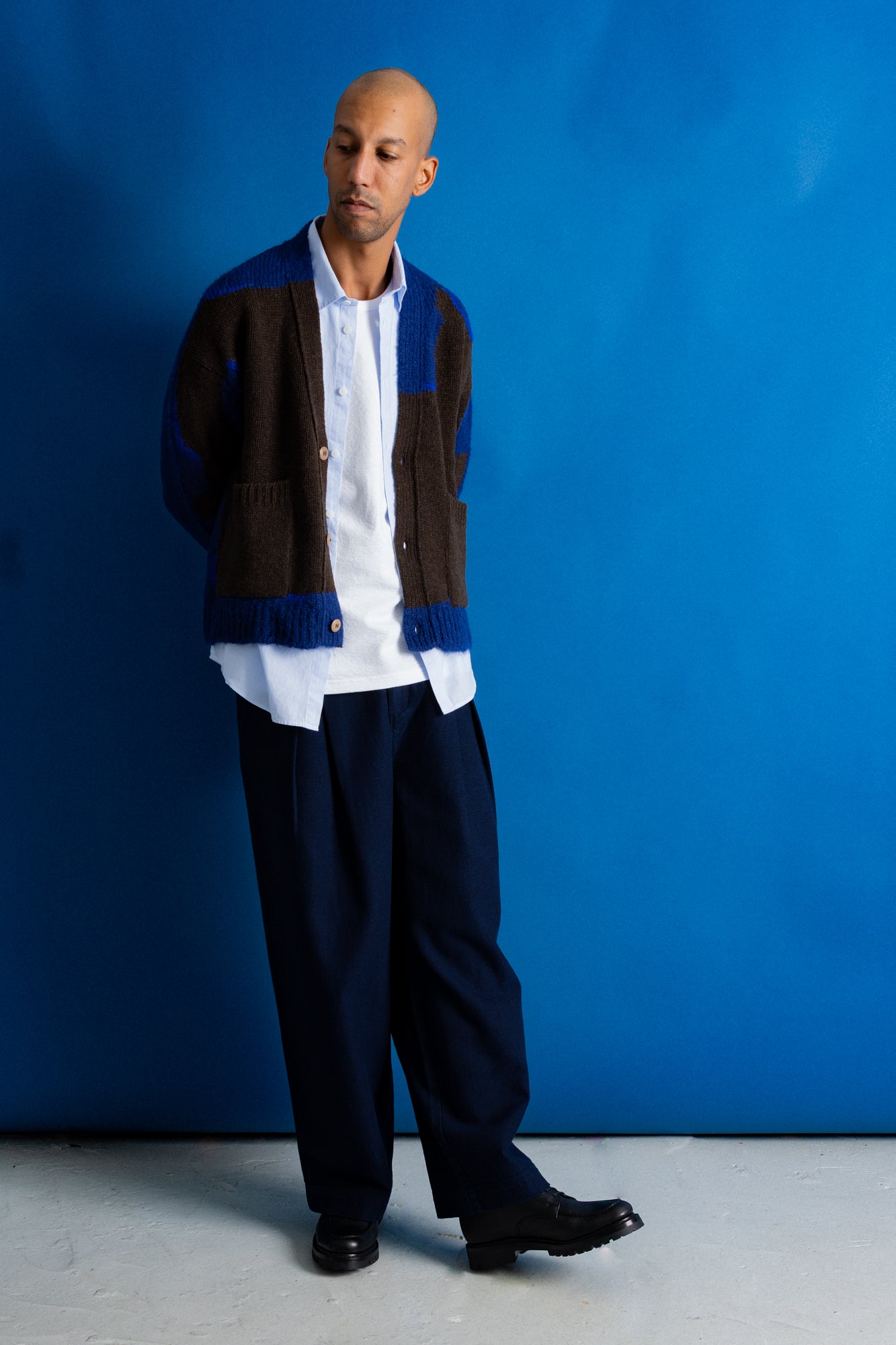 A man stands against a bright blue studio backdrop wearing a brown and blue color-block knit cardigan over a white button-up shirt, dark navy trousers, and black shoes.