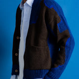 A man wearing a blue and brown color-blocked knit cardigan over a white collared shirt, standing against a solid blue background.