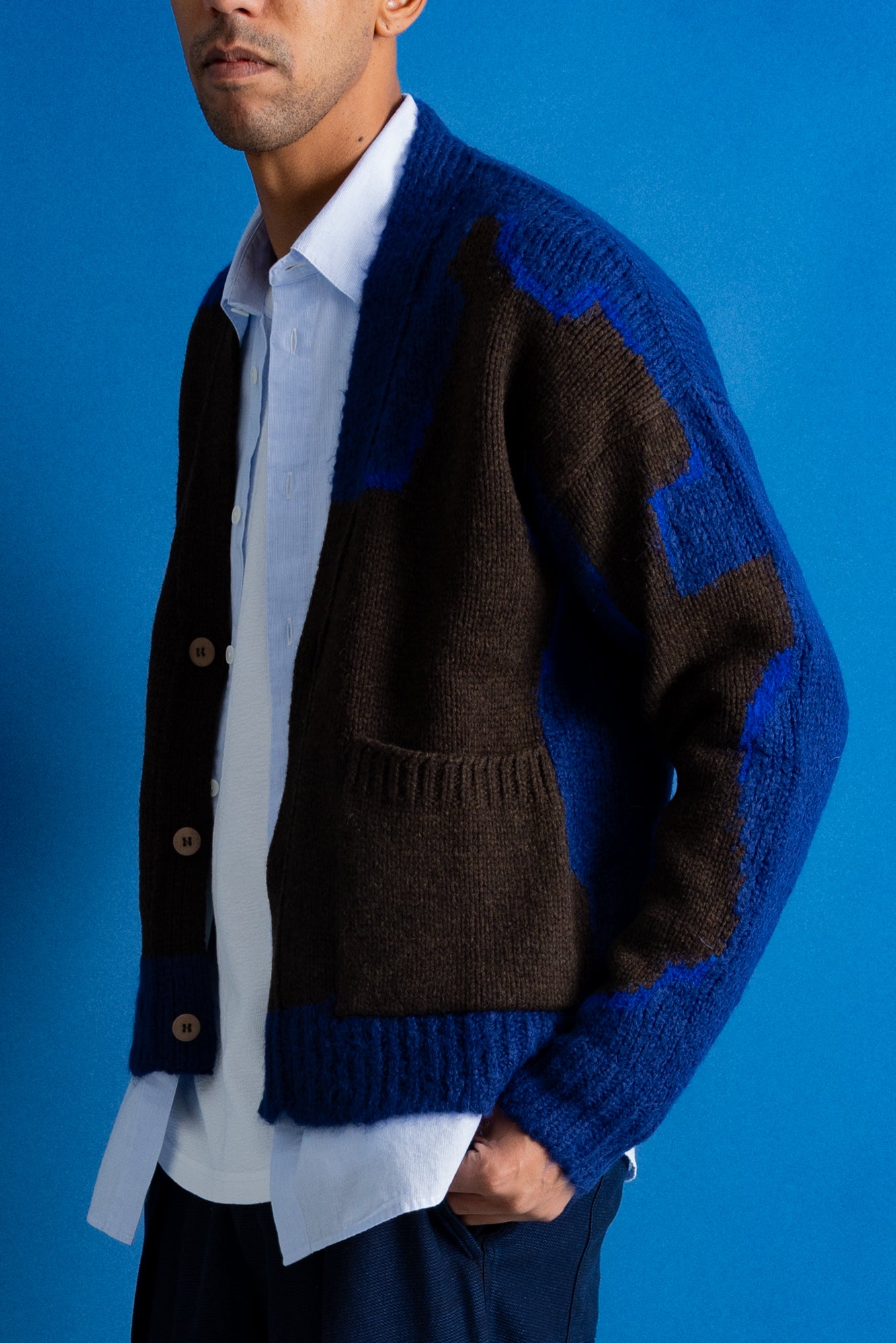 A man wearing a blue and brown color-blocked knit cardigan over a white collared shirt, standing against a solid blue background.