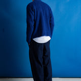 A man viewed from behind wearing a textured navy blue knit cardigan over a white shirt and dark trousers, standing against a solid blue backdrop.