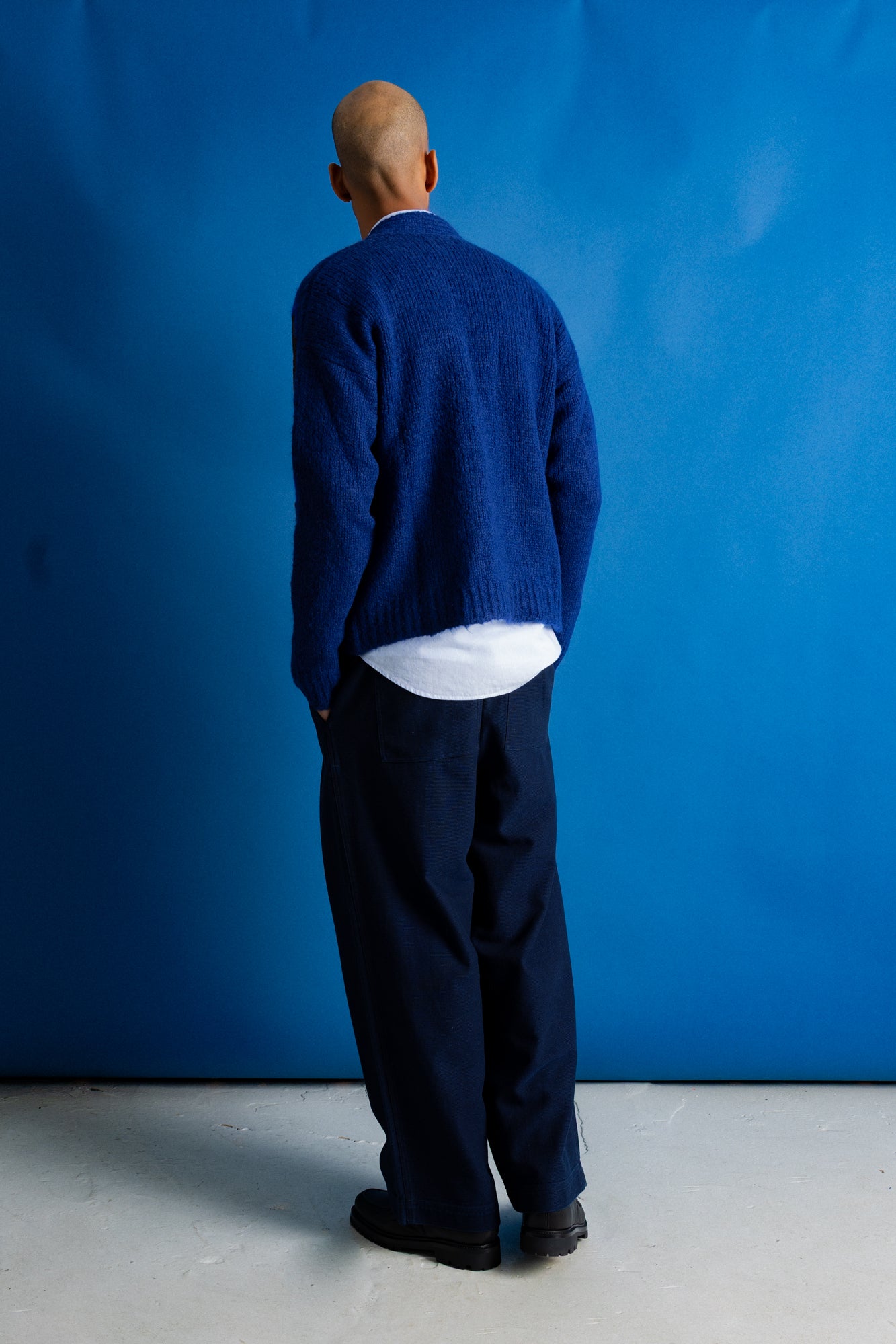 A man viewed from behind wearing a textured navy blue knit cardigan over a white shirt and dark trousers, standing against a solid blue backdrop.