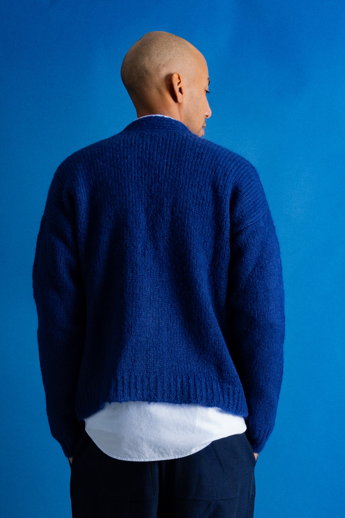 A man viewed from behind wearing a textured royal blue knit cardigan over a white shirt and dark trousers, set against a solid blue studio background.