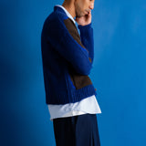 A man in a blue and brown knit cardigan over a white shirt and dark trousers, standing in profile against a solid blue background.