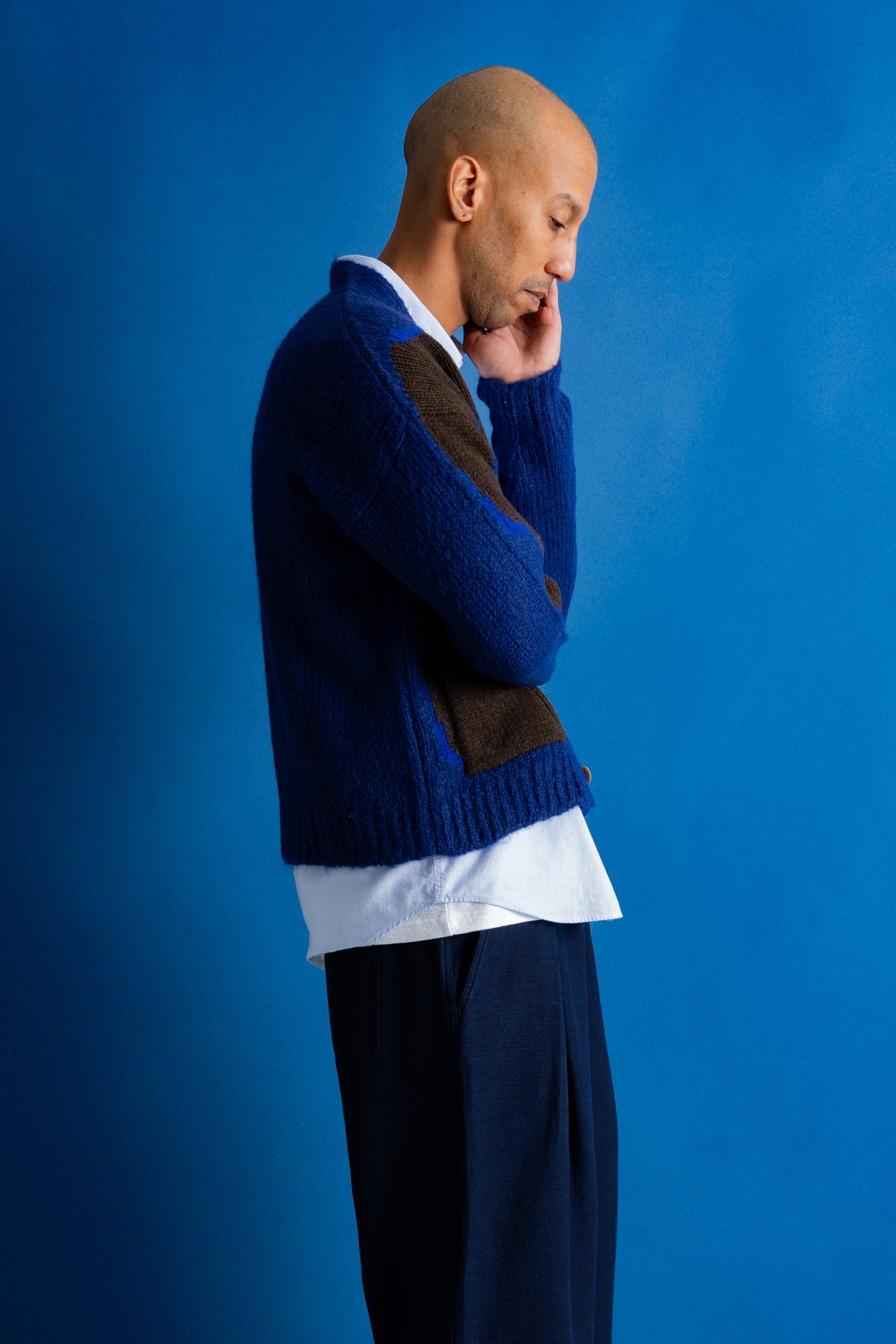 A man in a blue and brown knit cardigan over a white shirt and dark trousers, standing in profile against a solid blue background.