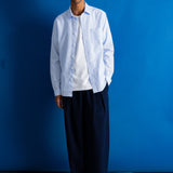 A man stands against a bright blue studio backdrop wearing a light blue button-down shirt over a white t-shirt, loose dark navy trousers, and black shoes.
