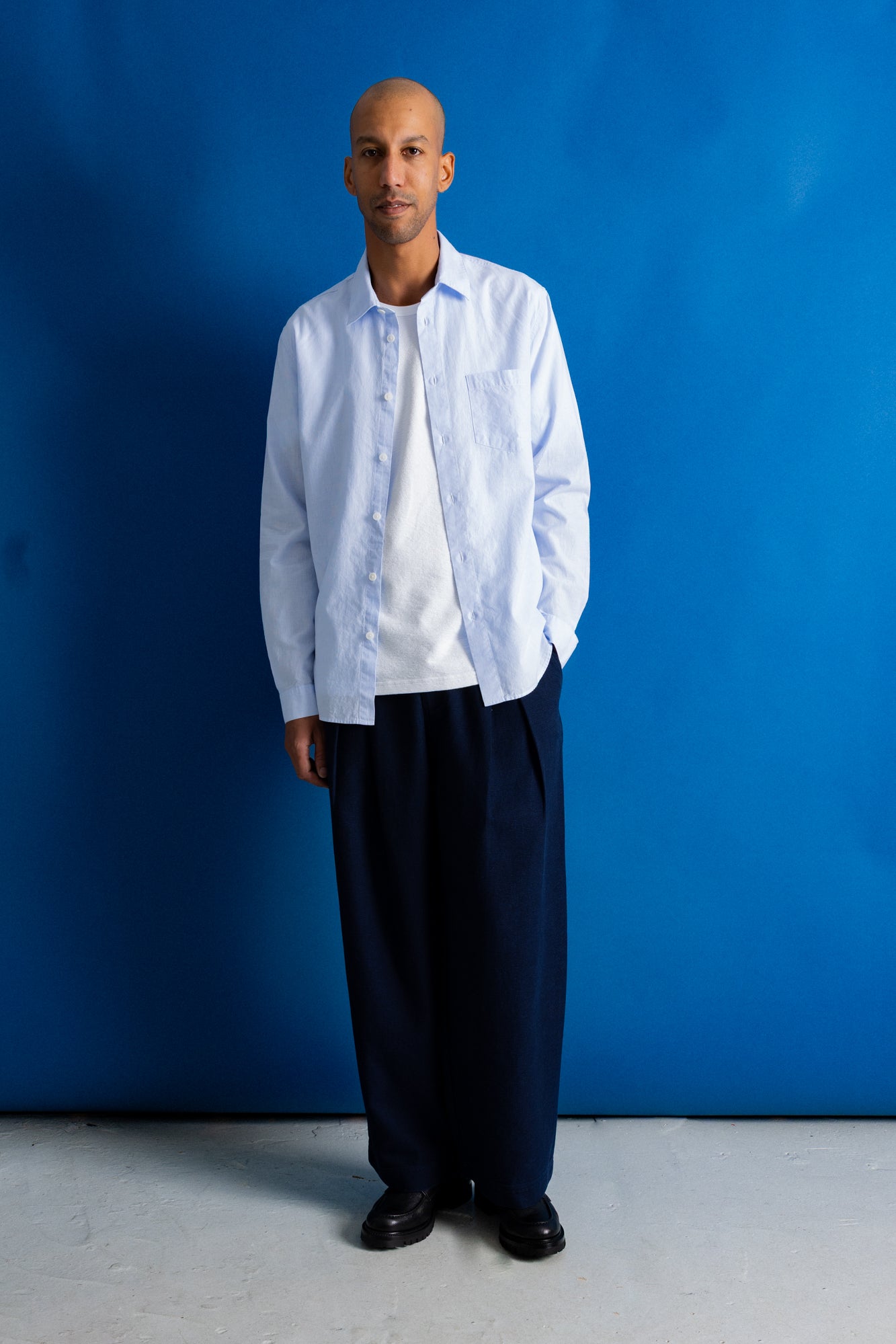 A man stands against a bright blue studio backdrop wearing a light blue button-down shirt over a white t-shirt, loose dark navy trousers, and black shoes.
