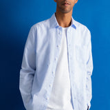 A man wearing a light blue long-sleeve button-down shirt over a white t-shirt and dark navy pants, standing against a solid blue background.