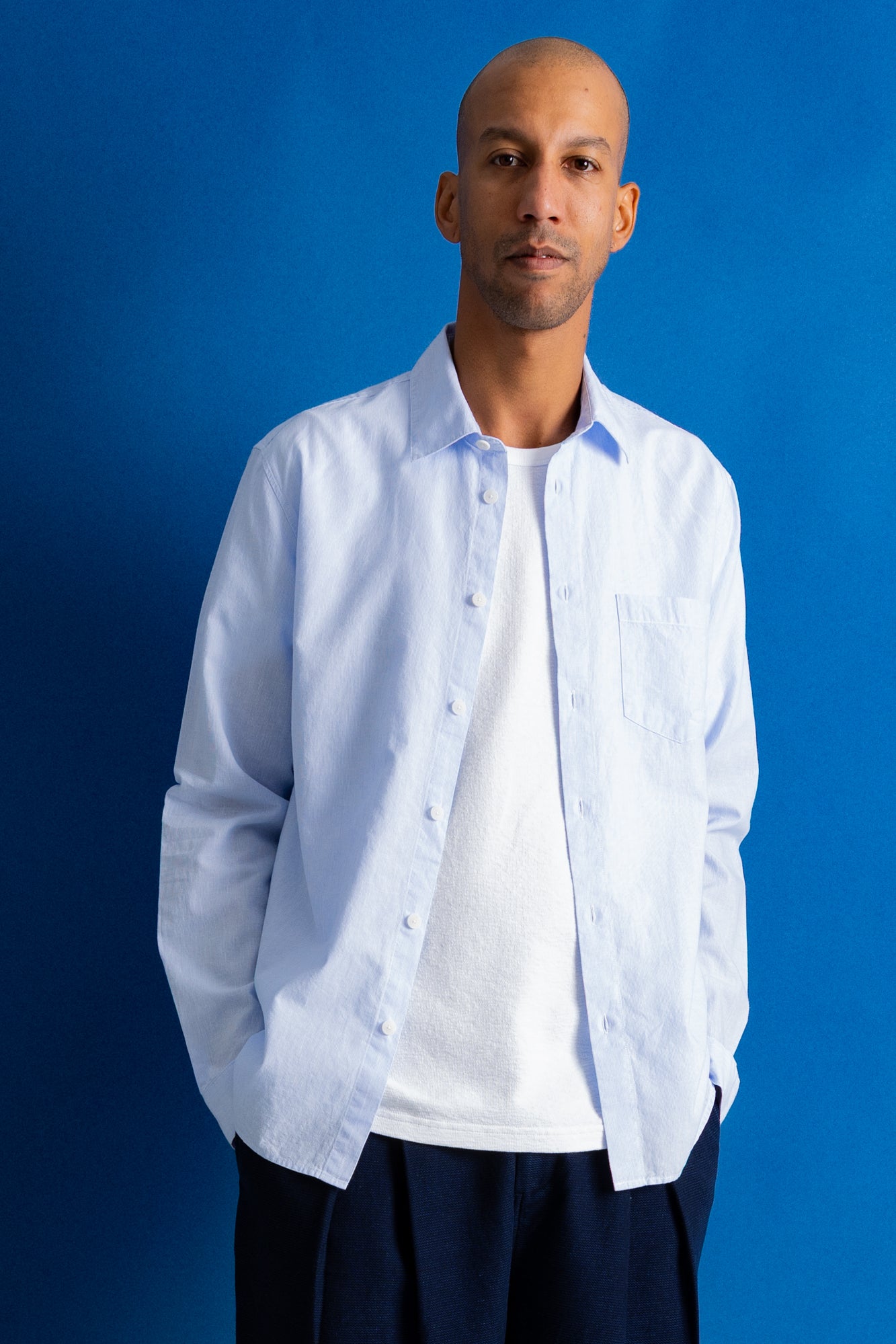 A man wearing a light blue long-sleeve button-down shirt over a white t-shirt and dark navy pants, standing against a solid blue background.