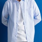 A man wearing a light blue long-sleeve button-up shirt over a white t-shirt, set against a solid blue studio background.