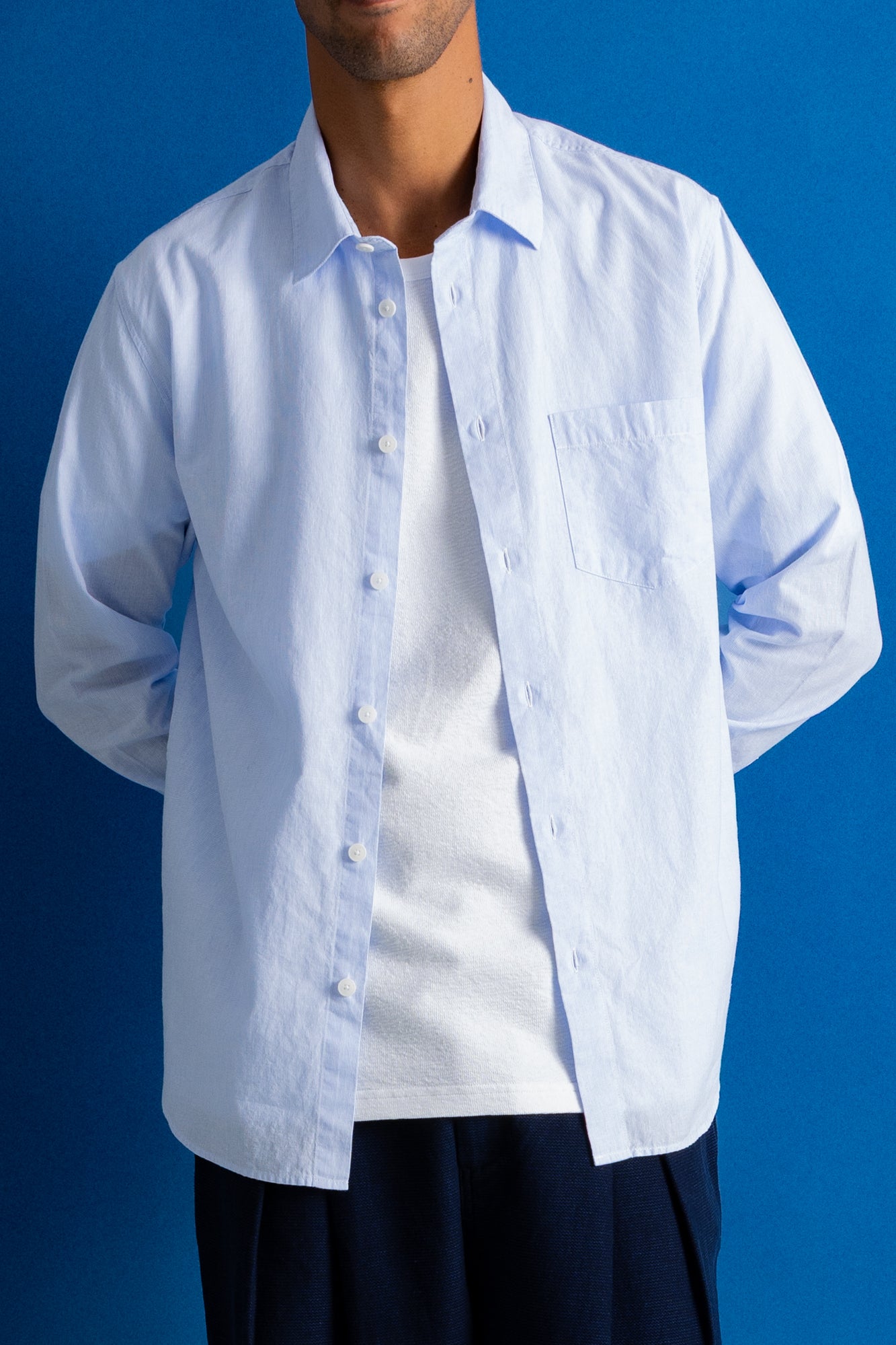 A man wearing a light blue long-sleeve button-up shirt over a white t-shirt, set against a solid blue studio background.