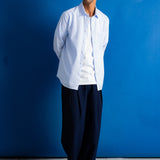 A man stands against a bright blue studio backdrop wearing a light blue button-up shirt over a white undershirt, loose dark navy trousers, and black shoes.