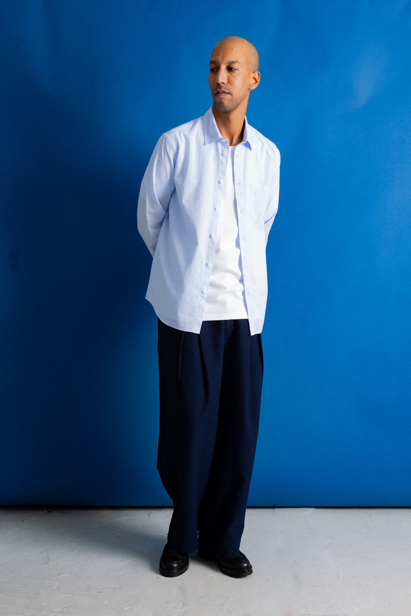 A man stands against a bright blue studio backdrop wearing a light blue button-up shirt over a white undershirt, loose dark navy trousers, and black shoes.