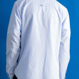 A man viewed from behind wearing a light blue long-sleeve button-down shirt and dark trousers, standing against a solid blue background.