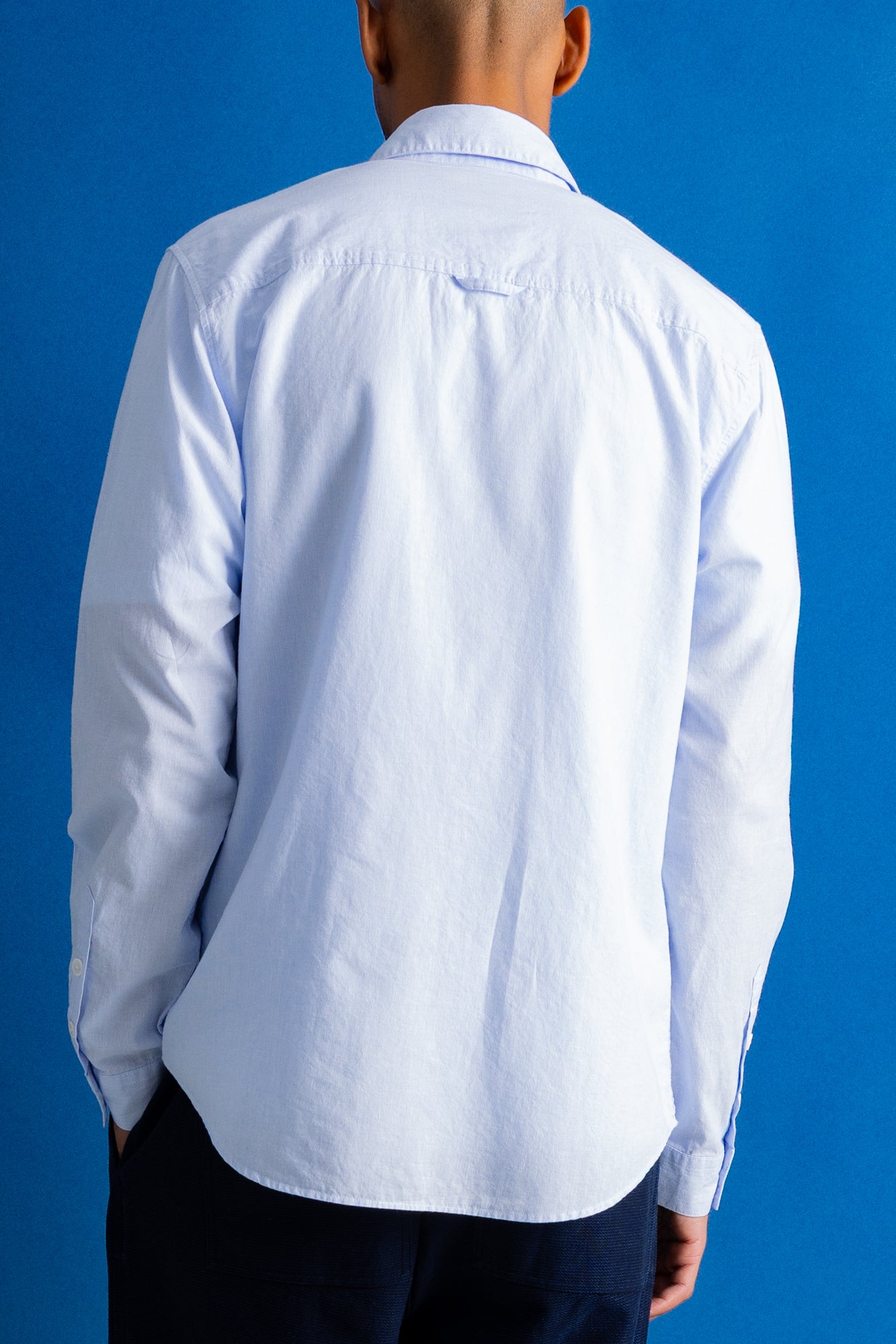 A man viewed from behind wearing a light blue long-sleeve button-down shirt and dark trousers, standing against a solid blue background.