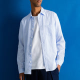 A man wearing a light blue long-sleeve button-down shirt over a white t-shirt and dark navy trousers, standing against a solid blue studio background.