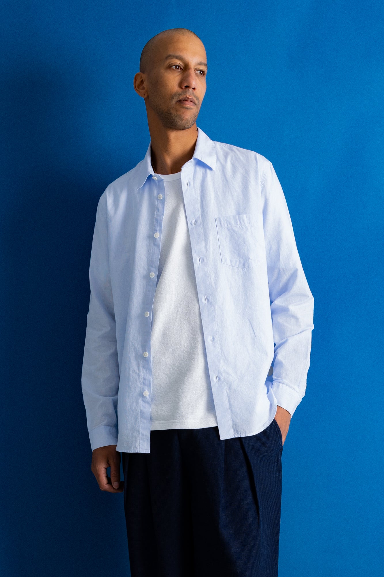 A man wearing a light blue long-sleeve button-down shirt over a white t-shirt and dark navy trousers, standing against a solid blue studio background.