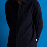 A man wearing a dark navy blue textured long-sleeve button-up shirt, shown from the chin down against a solid blue background.
