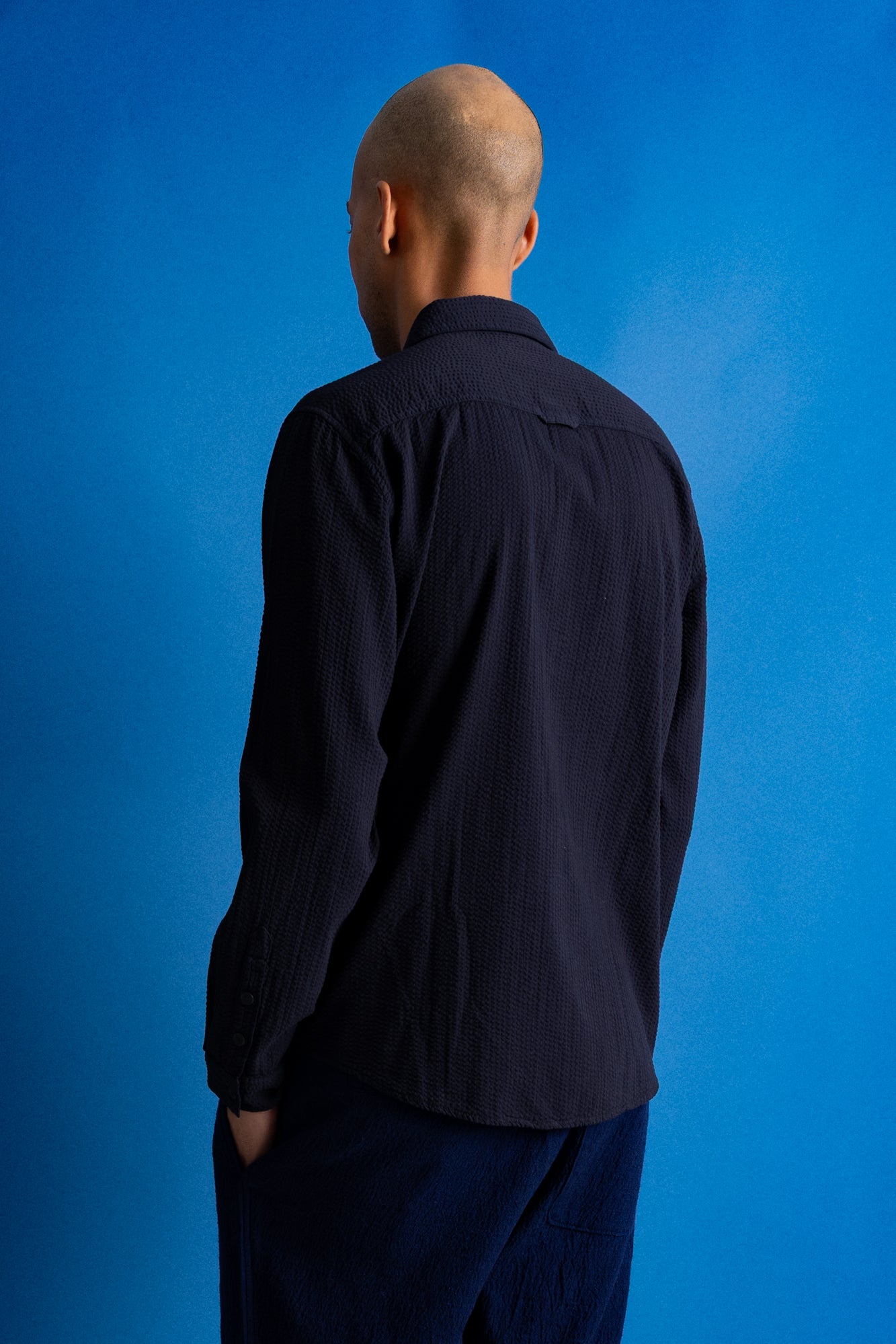 A man viewed from behind wearing a dark navy blue long-sleeve button-up shirt and matching dark trousers, standing against a vibrant blue studio background.