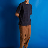 A man wearing a navy blue and black striped short-sleeve t-shirt, brown pants, and black sneakers, standing against a vibrant blue studio backdrop.