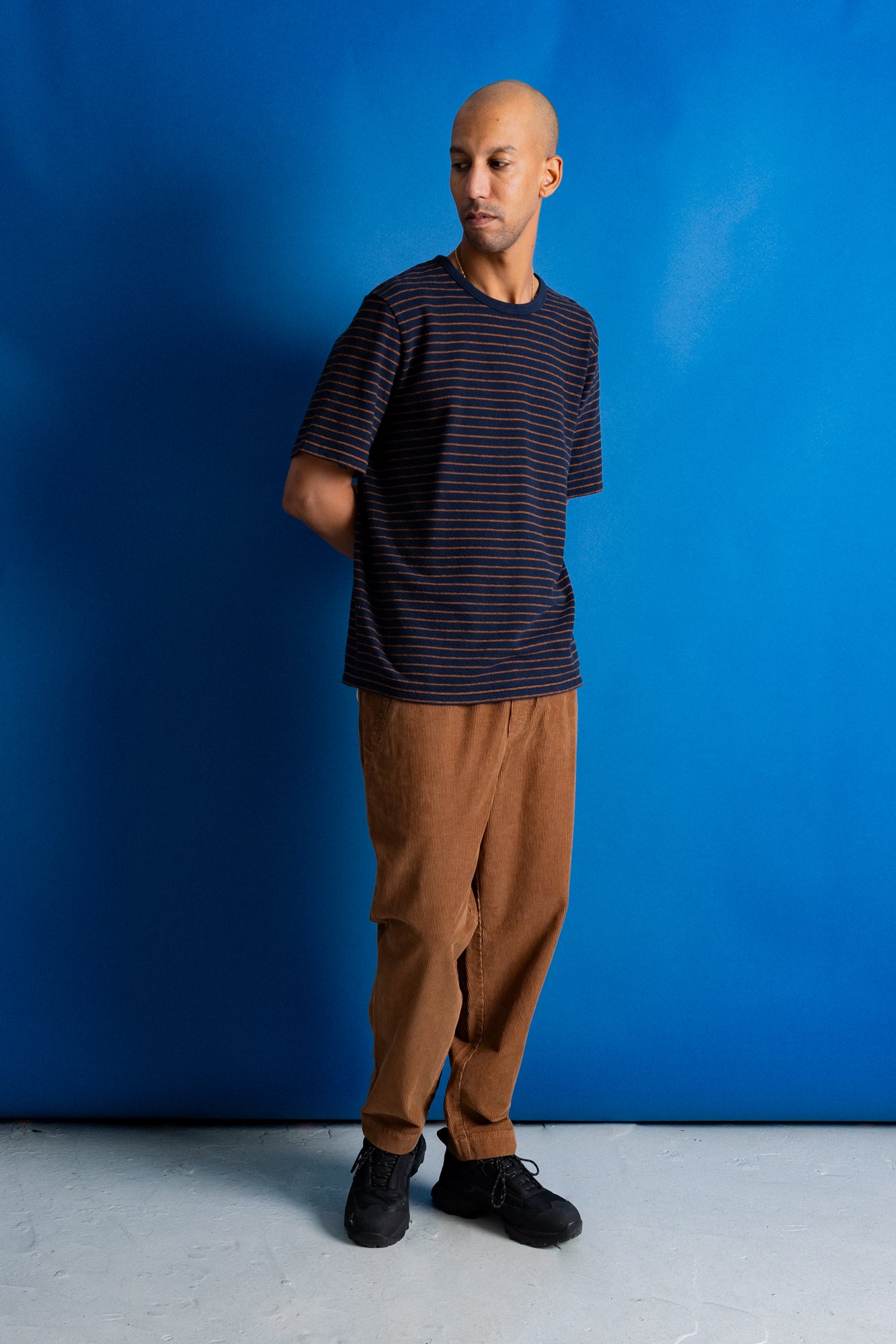 A man wearing a navy blue and black striped short-sleeve t-shirt, brown pants, and black sneakers, standing against a vibrant blue studio backdrop.