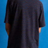 A person viewed from the back wearing a short-sleeve crew-neck t-shirt with thin horizontal navy and orange stripes, paired with brown pants, set against a solid blue background.