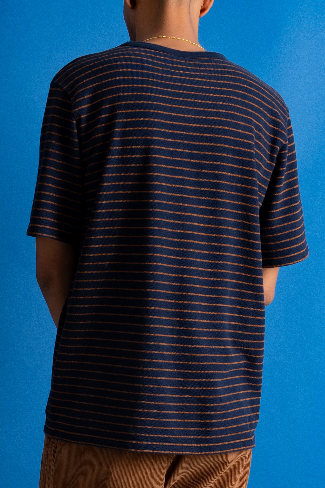 A person viewed from the back wearing a short-sleeve crew-neck t-shirt with thin horizontal navy and orange stripes, paired with brown pants, set against a solid blue background.