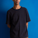 A man wearing a navy blue crewneck t-shirt with thin orange horizontal stripes, paired with brown corduroy pants, standing against a solid blue background.
