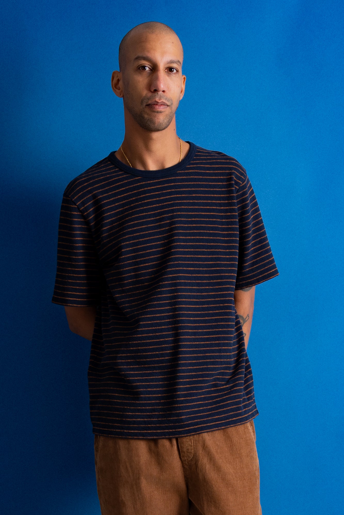 A man wearing a navy blue crewneck t-shirt with thin orange horizontal stripes, paired with brown corduroy pants, standing against a solid blue background.