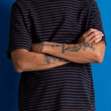 A man wearing a navy blue short-sleeve t-shirt with thin orange horizontal stripes, paired with brown pants, standing against a solid blue background with his arms crossed.