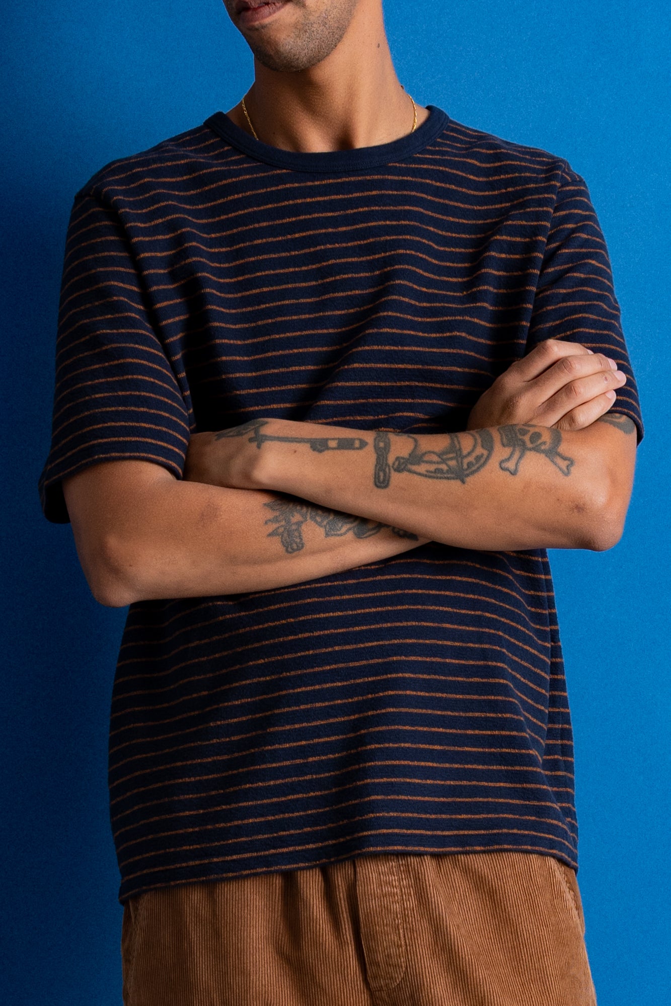 A man wearing a navy blue short-sleeve t-shirt with thin orange horizontal stripes, paired with brown pants, standing against a solid blue background with his arms crossed.