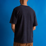 A man viewed from behind wearing a navy blue short-sleeve t-shirt with thin orange horizontal stripes and brown pants, standing against a solid blue background.