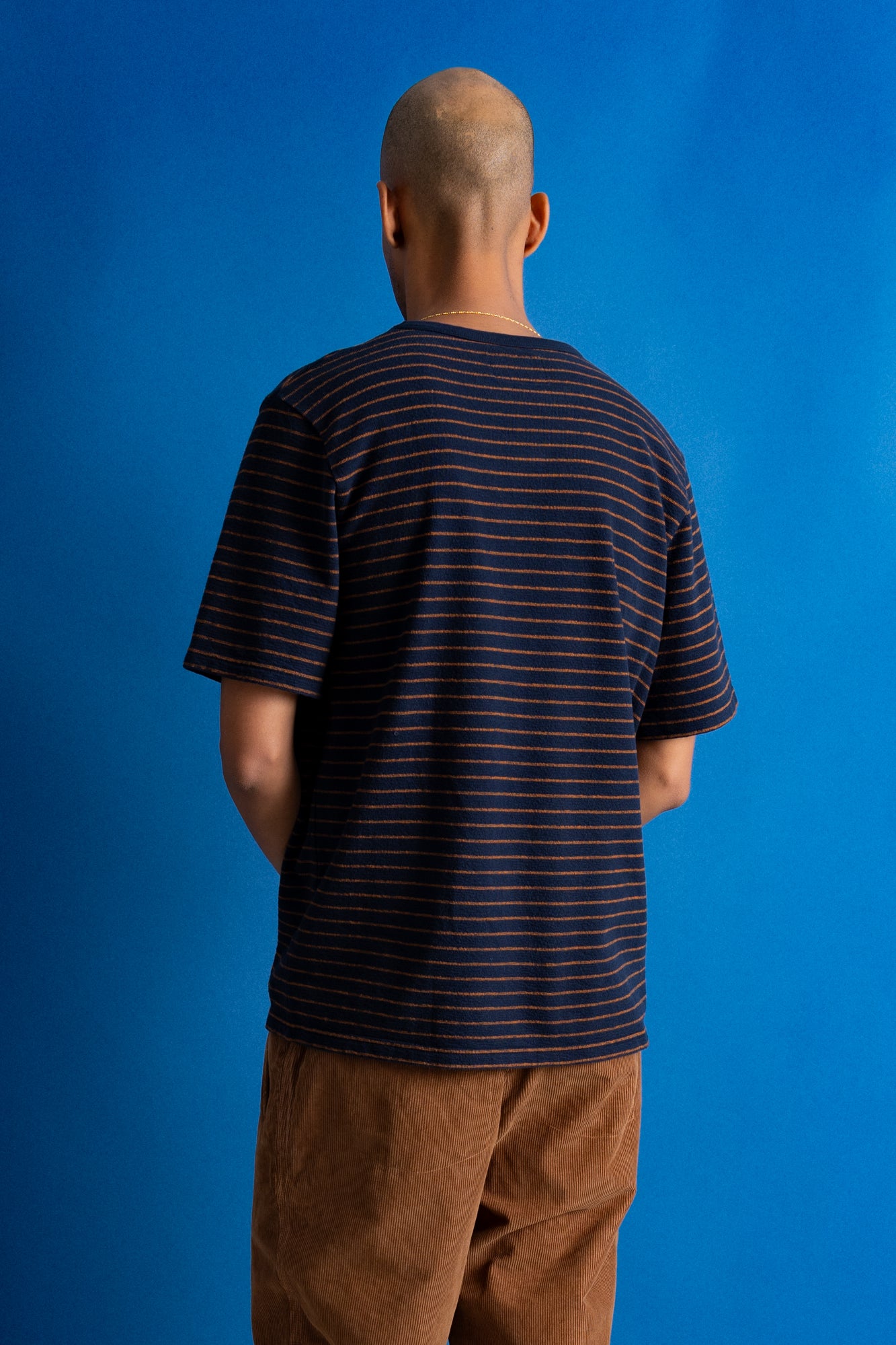 A man viewed from behind wearing a navy blue short-sleeve t-shirt with thin orange horizontal stripes and brown pants, standing against a solid blue background.