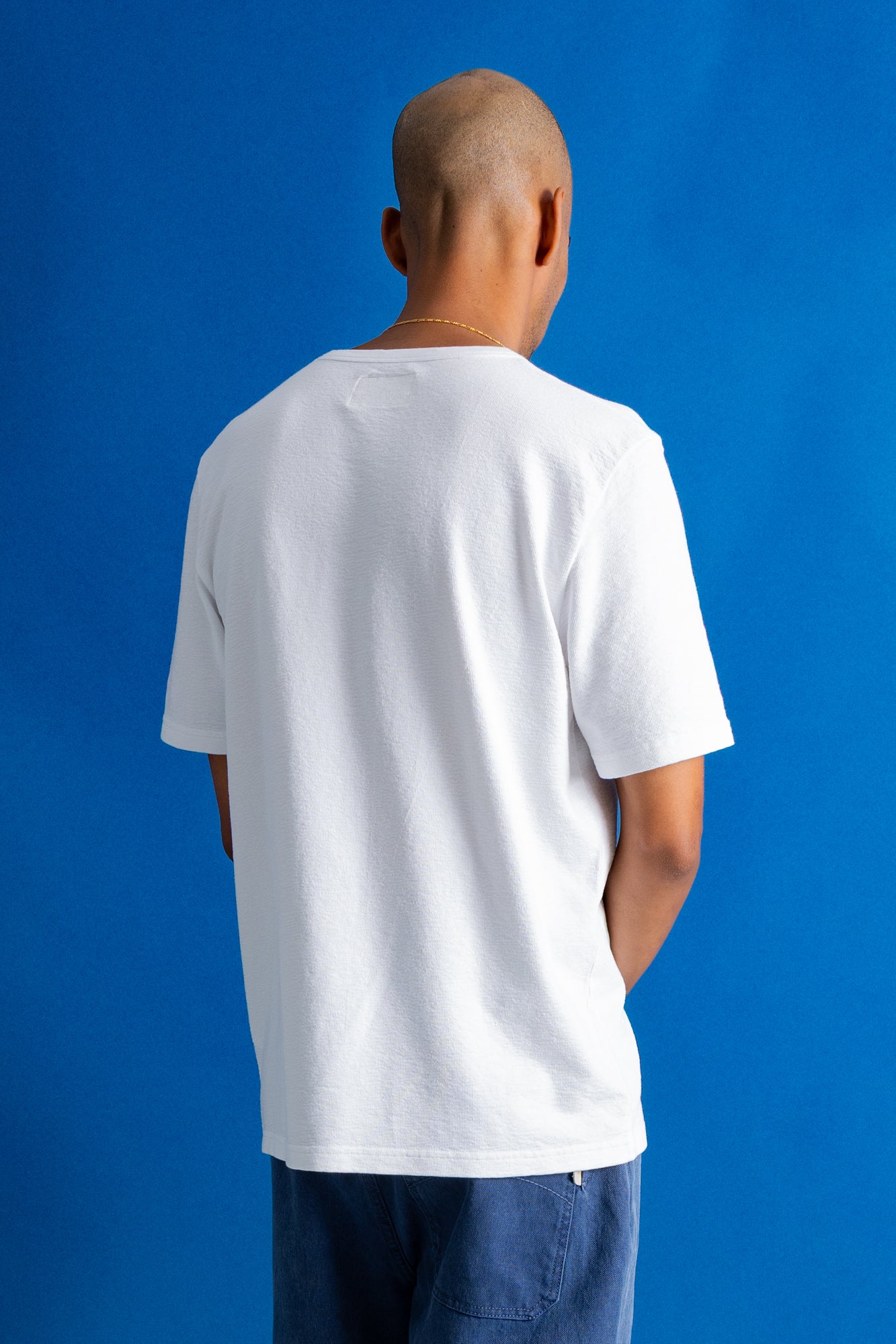 A man viewed from behind wearing a plain white short-sleeve crewneck t-shirt and blue jeans, standing against a solid blue studio background.